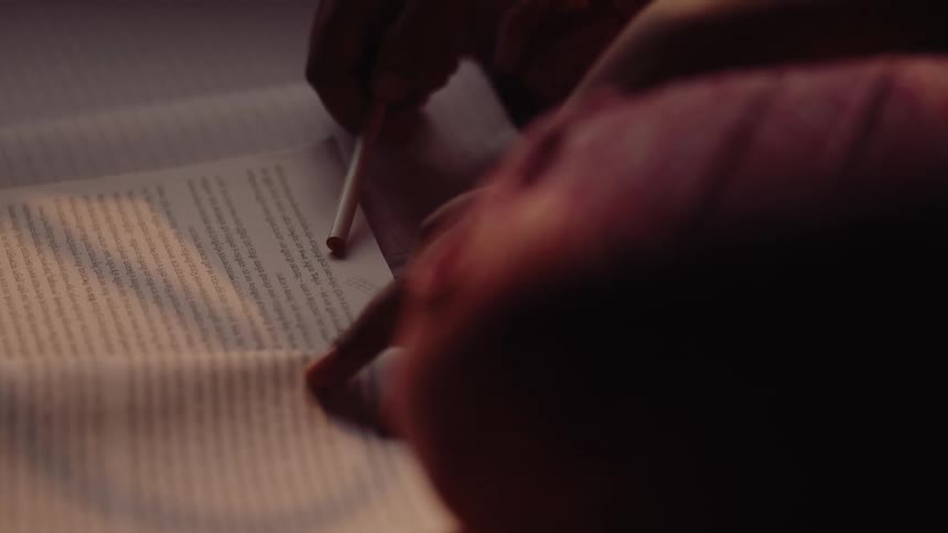 Someone reading a book with a pen in hand in a dark room at sunset