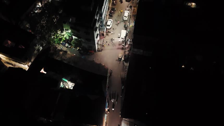A nighttime aerial shot captures a quiet dimly lit urban neighborhood street with sparse foot traffic and streetlights casting long shadows