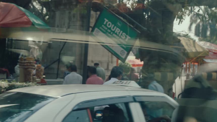 A lively urban street scene shows pedestrians market stalls and vehicles moving past a historic monument in a bustling south asian city during midday