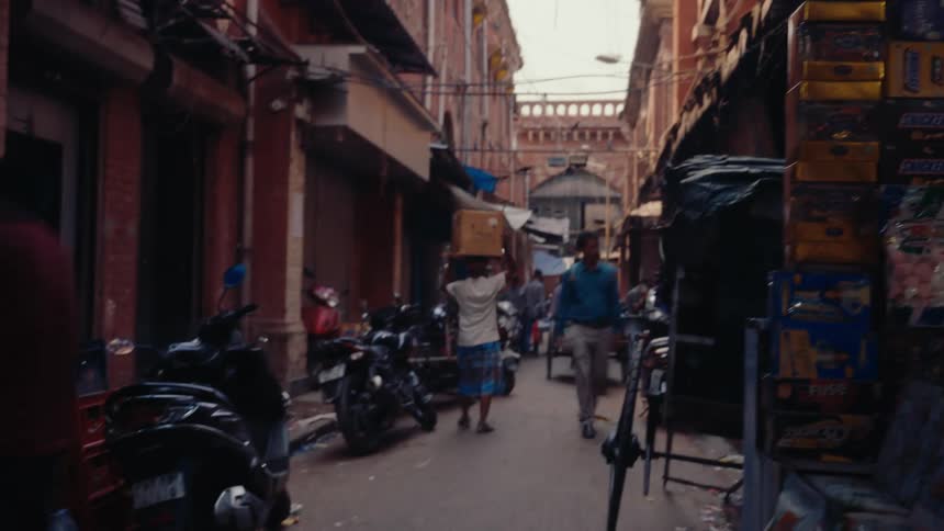 A man wearing a blue shirt walks along a narrow street at midday passing another person carrying a box on his head with a row of scooters parked along the roadside