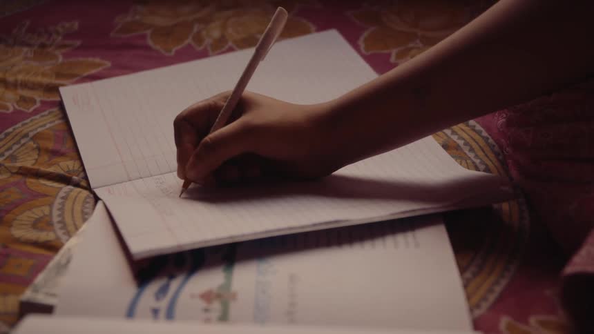 A hand is intently writing in a notebook resting on a patterned bedspread illuminated by a warm light source
