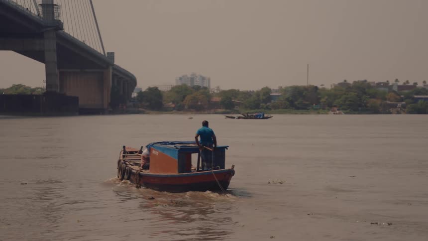 Two men ride a small wooden boat across a calm river with a large urban bridge and cityscape in the background