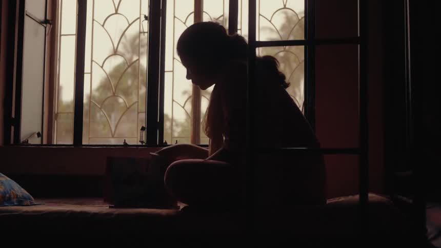 A young woman sits crosslegged in silhouette by a window reading or writing in a dimly lit room