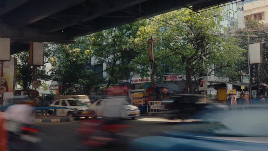 Moving vehicles cross an urban avenue under a viaduct with trees and businesses in the background