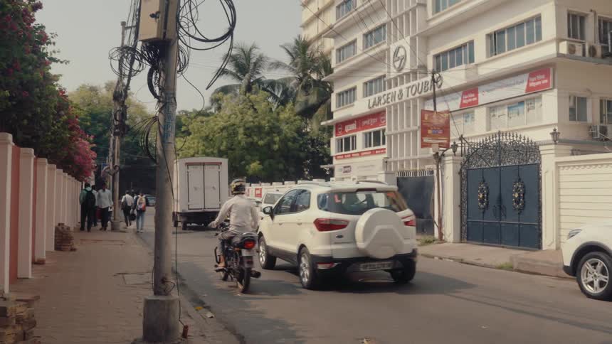 A busy urban street scene shows various vehicles and pedestrians moving past commercial and residential buildings in broad daylight