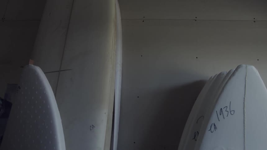 Unfinished surfboards rest against a wall in a warehouse during daylight