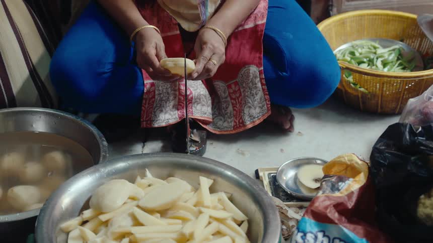 A woman is seated on the floor peeling and cutting potatoes in a kitchenlike setting surrounded by bowls of vegetables and cooking materials