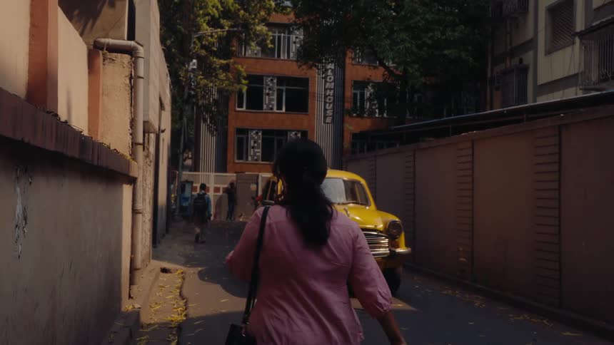 A yellow taxi drives down a narrow alleyway lined with buildings while several pedestrians walk through the urban street scene