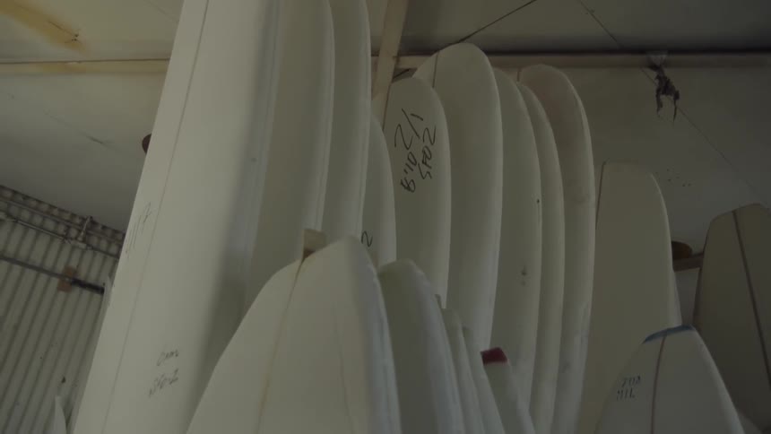 Unfinished surfboards line the interior of a warehouse during the day