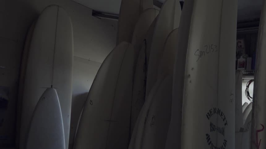 Unfinished surfboards are stacked against a wall