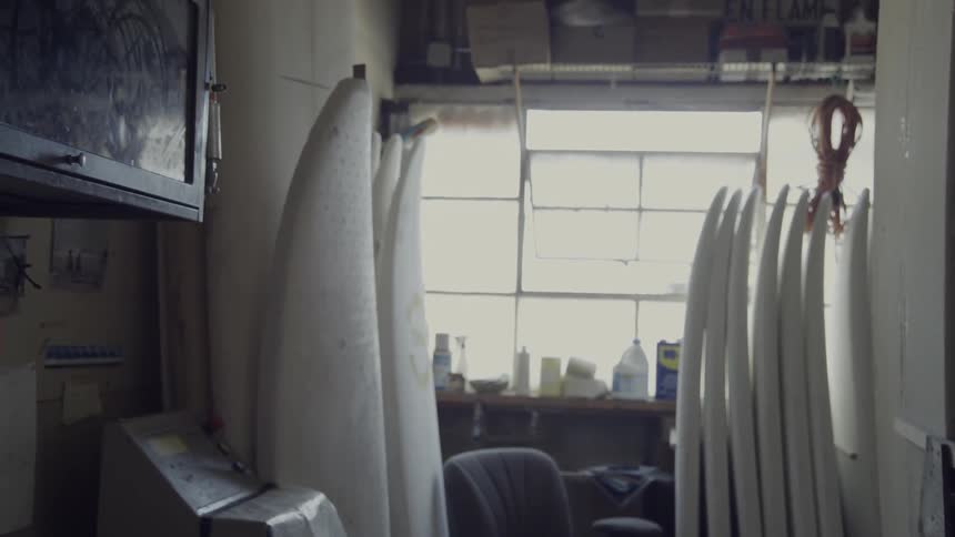Unfinished surfboards are stacked against a wall