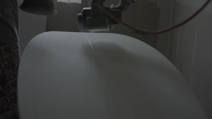 A man runs a tool over an unfinished surfboard