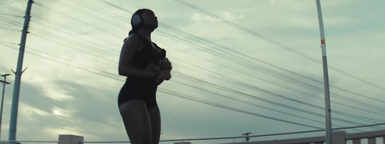 A woman wearing workout gear and headphones exercises outdoors on an urban rooftop at dusk