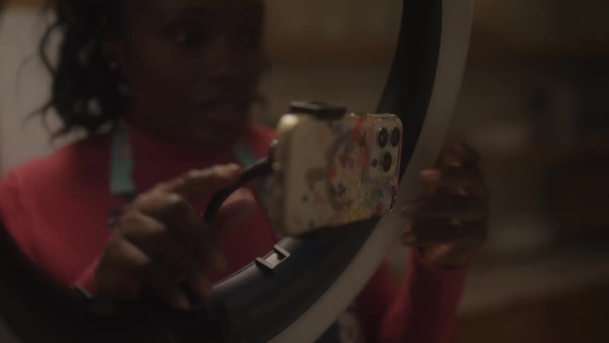 A woman in a kitchen is recording herself with a phone through a ring light possibly filming content or taking a selfie