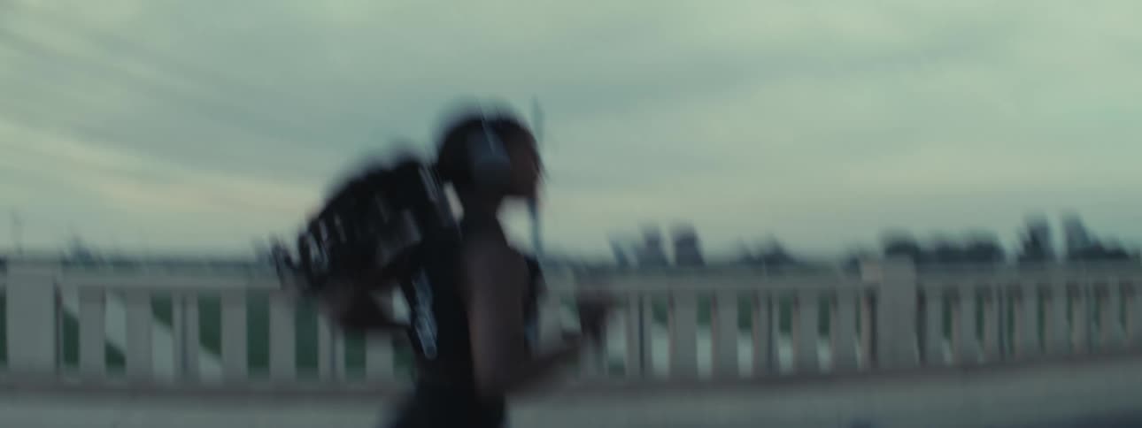 A woman in athletic wear runs across a bridge wearing headphones with a city skyline visible in the background