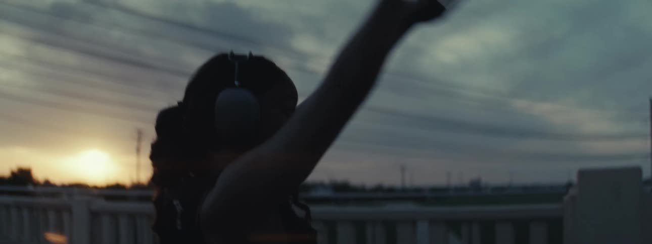 A young black woman stretching by the roadside at sundown wearing headphones with sunlight still visible and a series of power lines in the background