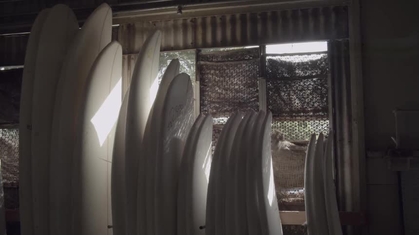 Unfinished surfboards are stacked against a wall