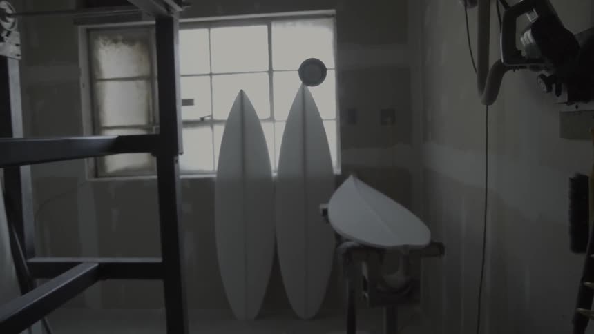 An unfinished surfboard rests on a workbench in a workshop during the day
