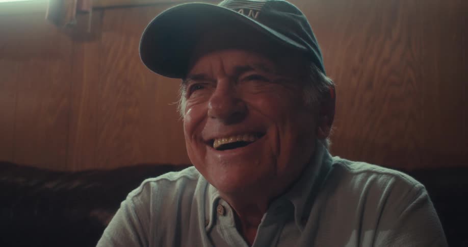 An elderly man wearing a hat labeled veteran sits in his living room at midday laughing as he watches something on his mobile device