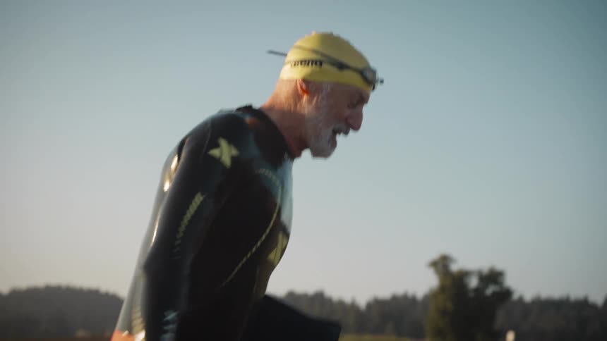 Older man wearing a wet suit and swimming cap smiles and talks while walking through a field during the daytime