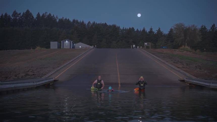 Three people are wading in water at a boat ramp under a dusky sky with the moon visible in the background