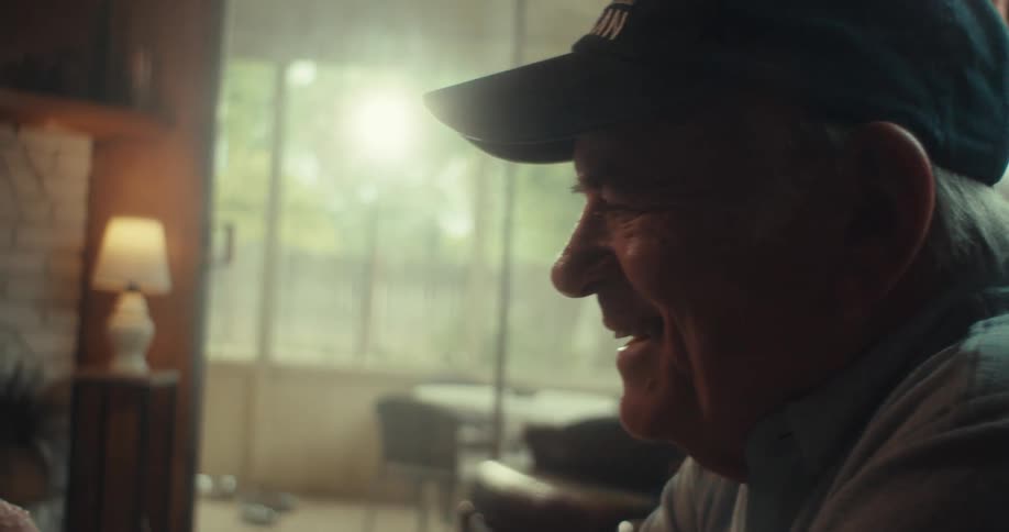 An elderly man is sitting indoors in a cozy softly lit room using a smartphone with a thoughtful expression