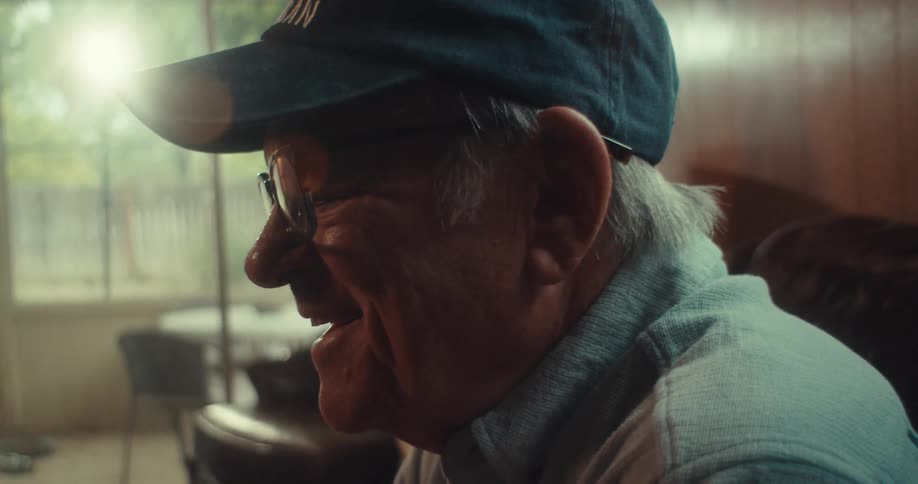 An elderly man wearing a hat and glasses sits in his living room at midday laughing while looking at a screen in front of him