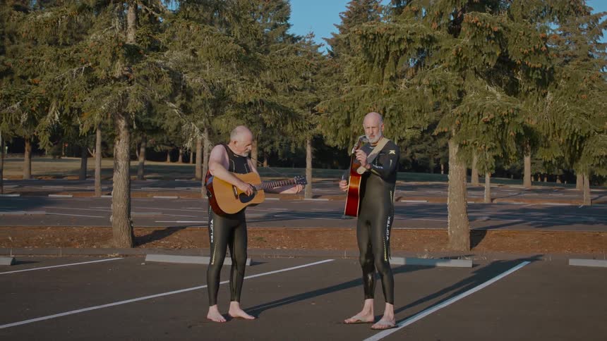 Two men in wetsuits play acoustic guitars barefoot in an empty parking lot surrounded by tall evergreen trees during golden hour