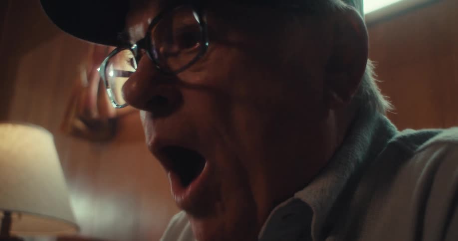 An elderly man wearing glasses and a cap is sitting indoors intently looking at a tablet in a warmly lit room