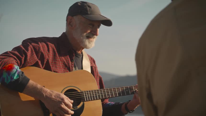 An elderly man plays an acoustic guitar outdoors while smiling warmly at someone offcamera suggesting a peaceful and joyful moment in nature