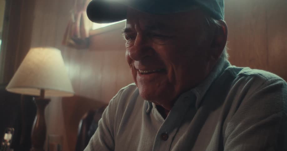 An elderly man wearing a veteran baseball hat sits in his living room at midday laughing while looking at a smartphone in his hand