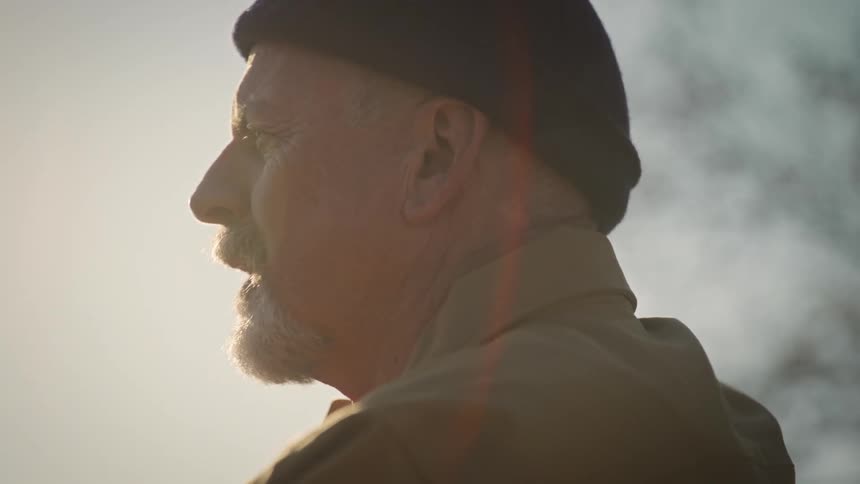 A contemplative elderly man in a beanie is seen in profile softly backlit as he speaks to someone just out of frame evoking a quiet reflective mood