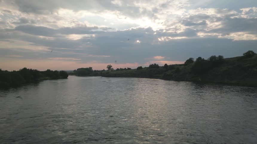 Birds fly over a river passing between the woods and hills while red tints the cloudy sky
