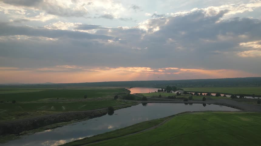 A serene landscape features a reflective pond in the midst of lush green fields under a cloudfilled sky at sunset