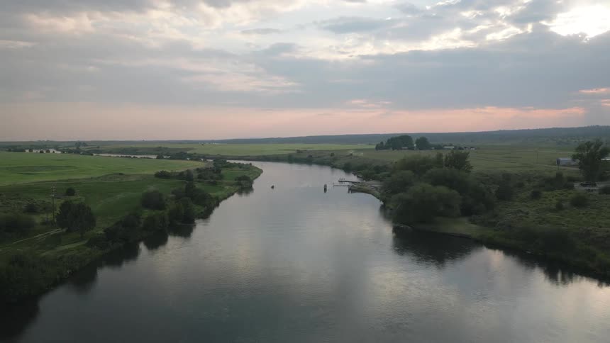 A wide aerial view captures a calm river flowing through green fields beneath a dramatic sunrise or sunset sky while birds fly by