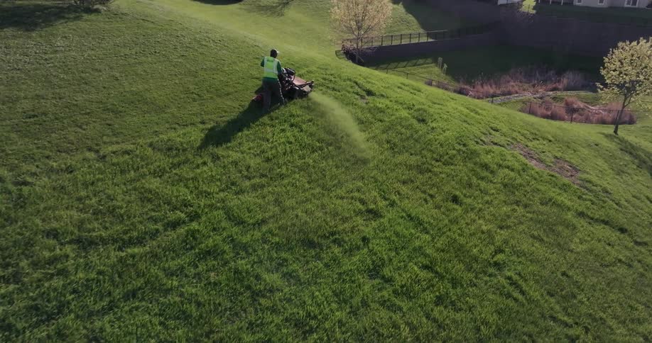 Aerial view of a gardener operating a machine and cutting grass on a hillside