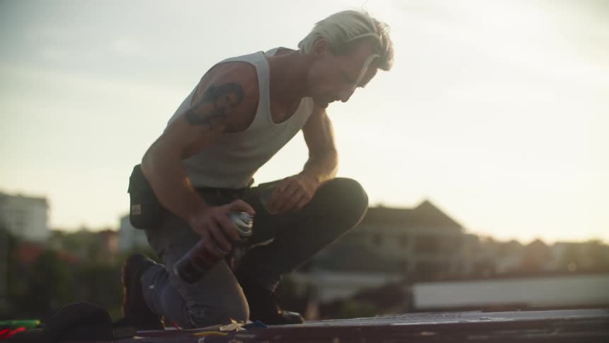 A person wearing a lightcolored tank top kneels on a rooftop applying spray paint to a surface at evening