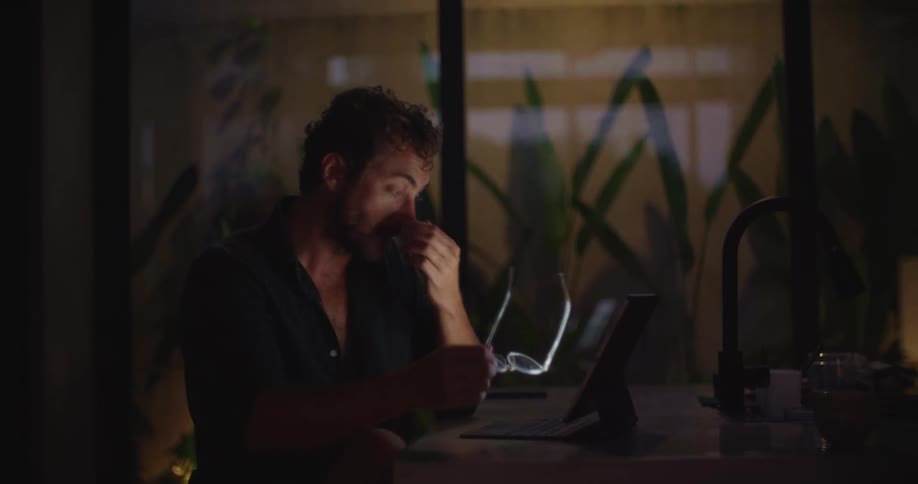 A man works late at a dimly lit kitchen counter focused on his laptop before growing visibly tired and rubbing his eyes