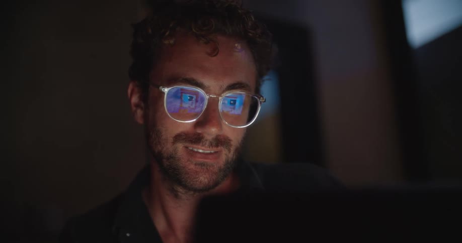Man with curly hair and glasses smiles at a screen reflections visible in his lenses in a dimly lit room