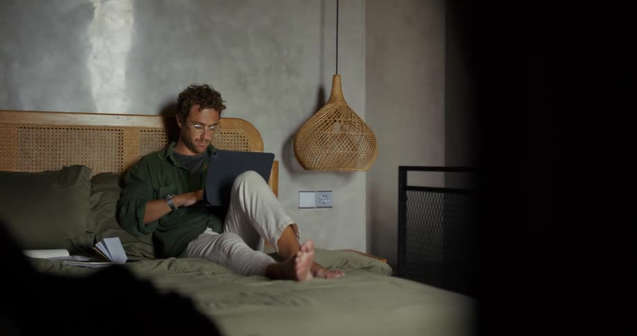 A man wearing glasses sits on his bed with his laptop on his lap operating it