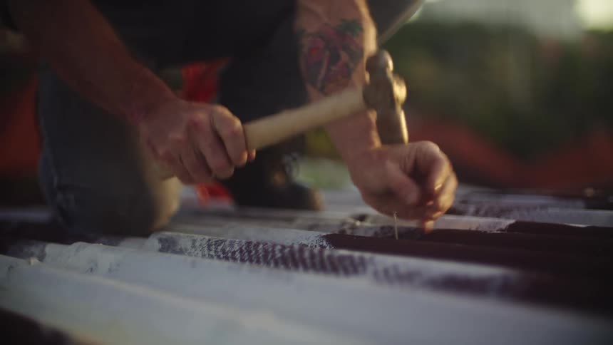 A person is shown on their knees hammering a nail into a corrugated surface likely a roof
