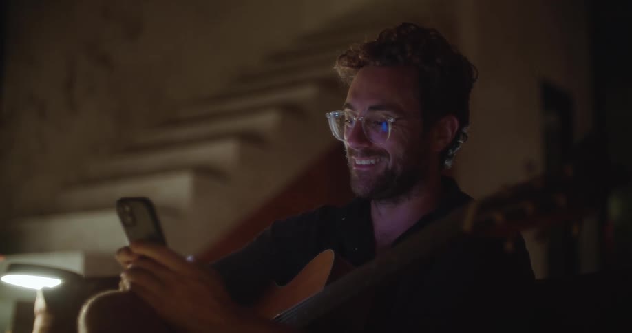 Man smiles as he texts on a phone while holding a guitar