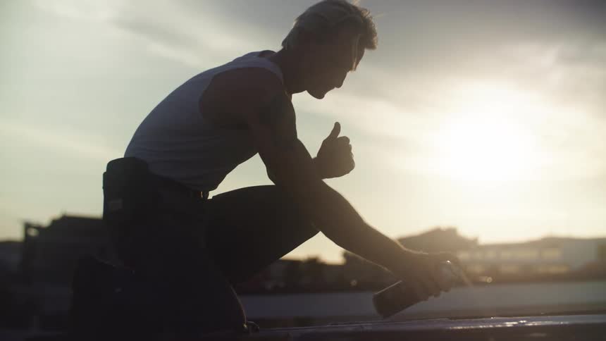 A person wearing a lightcolored tank top kneels on a rooftop applying spray paint to a surface