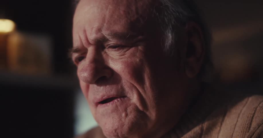 Closeup of an elderly mans face in a dimly lit room showing alternating expressions of happiness sadness and deep thought