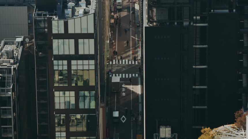 A topdown shot shows a narrow city street flanked by tall buildings with pedestrians crossing a zebra crossing