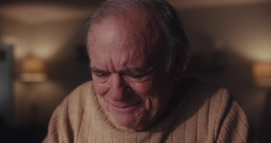 An elderly man sits indoors smiling warmly while video chatting on a laptop in a cozy softly lit room