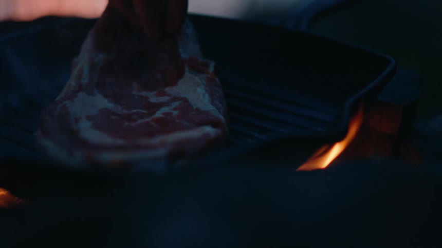 An adult cooks a steak on an outdoor grill at night