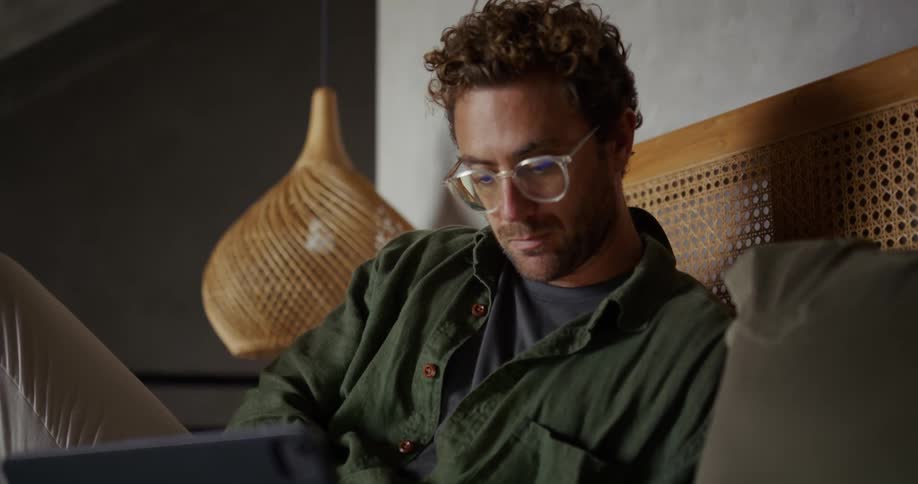 A casually dressed man with glasses sits indoors on a couch focused intently on a screen possibly a tablet or laptop