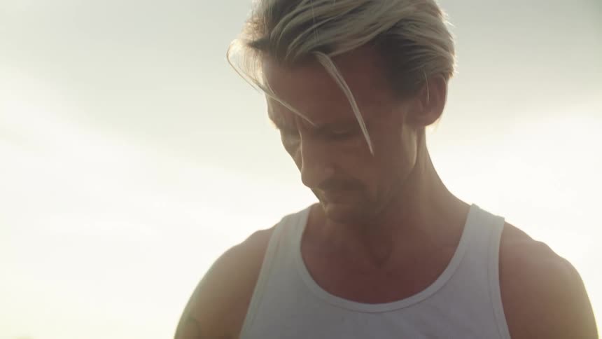 A man in a white tank top coils cables on a rooftop at sunset shown in closeup and medium shots highlighting his focus and muscular arms