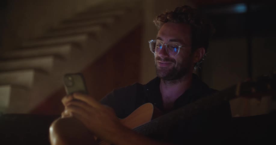 A man with glasses is sitting indoors in low light holding a smartphone while resting a guitar on his lap